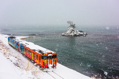 A colorful Japanese train moves through a snowy landscape near the ocean in Noto.