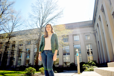 Sarah Milholland stands in front of an MIT building on a sunny day spring day. Leaves on the trees behind her are just beginning to emerge.
