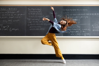 Janabel Xia dancing in front of a blackboard. Her back is arched, head thrown back, hair flying, and arms in the air as she looks at the camera and smiles.