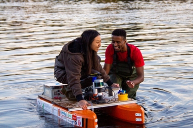 Audrey Chen crouches on top of a small orange boat while Jared Byars stands in the water, steadying the boat. Both are laughing.