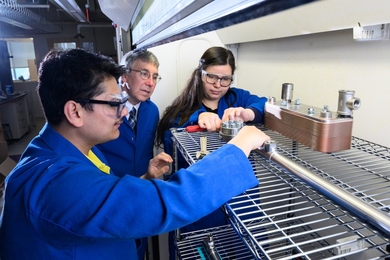 Three researchers in blue lab coats assemble a lab experiment on wire shelving 