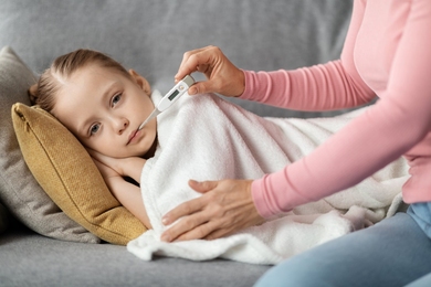 A little girl lies on a couch under a blanket while a woman holds a thermometer to the girl's mouth.