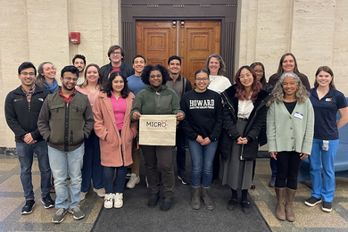 17 people pose outside a building with a brown door. Most wear jackets, and one holds a sign bearing the MICRO logo.