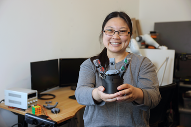 Sandra Liu poses for the camera holding her GelPalm prototype, a robotic hand with sensors. She is in a lab workspace with two computer monitors, a Rubik's cube, and electronic equipment.