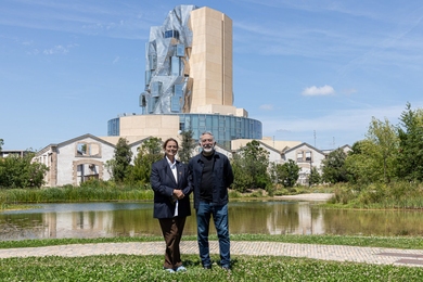 Maja Hoffmann and Hashim Sarkis pose outdoors on a sunny day. Behind them are a small pond, several older single-storey buildings, and a multi-storey building with a central tower, half metallic with windows jutting out in odd angles, and half tan stone