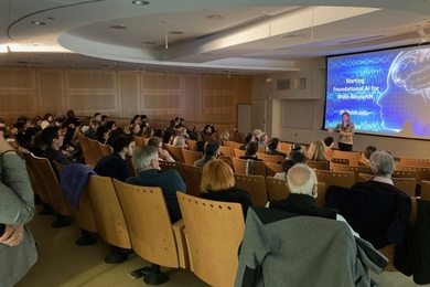 A woman speaks to a packed room of people. A large screen behind her displays a blue slide with the words, "Starting foundational AI for brain research."