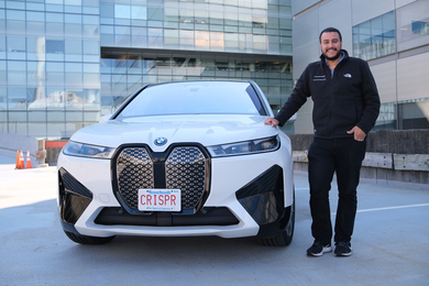 Omar Abudayyeh stands on a rooftop next to a white BMW SUV with the license plate, “CRISPR.” MIT lab buildings are in the background.