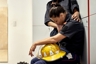 A woman in uniform sitting in a locker room with helmet in lap, head down, and eyes closed, being consoled by a colleague.