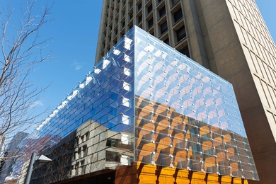 Exterior of the two-story glass Building 55 atrium extends from the concrete 21-story Building 54 tower, reflecting the sky and trees.