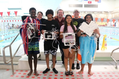 Seven people pose poolside. The four in front hold remotely operated underwater vehicles that they built.