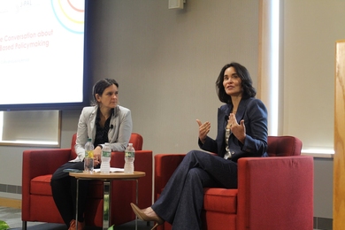 Laura Arnold and Esther Duflo sit in a conference space. Arnold gestures and speaks as Duflo looks at her.