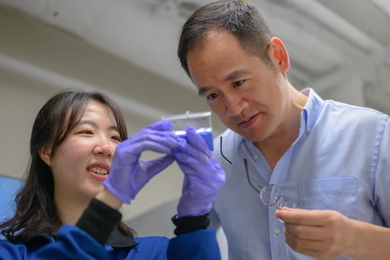 So Yeon Kim, wearing lab gear, holds up an out-of-focus object in her gloved hands for Ju Li's inspection