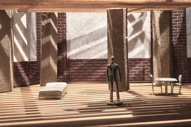 A small model shows a wooden man in a sparse room, with dramatic lighting from the windows.