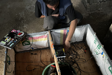 Ahmad Zakka squats by a wooden box that has been outfited with insulation and a bunch of wires