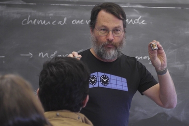 David Trumper stands in front of a chalkboard, holding up a small cylindrical electric motor in each hand