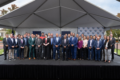 36 individuals pose on an outdoor stage under a pyramidal awning