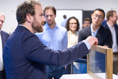 Aaron Baskerville-Bridges, shown in profile, shows a prototype window to several out-of-focus people standing to his left