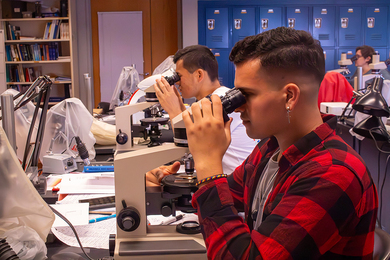 A student looks through a binocular microscope. Behind him, another student does the same.