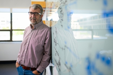 Curtis Smith leans on a glass whiteboard in an office with windows in the background