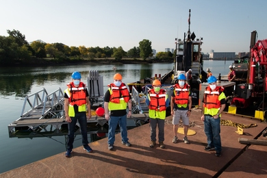 Stephen Murray, Jason Valenzano, David Kindler, Paul Ryu, and Andrew March, all in face masks, hard hats, and orange flotation vests, pose in front of a large metal frame floating in a waterway with Boston skyscrapers in the distance