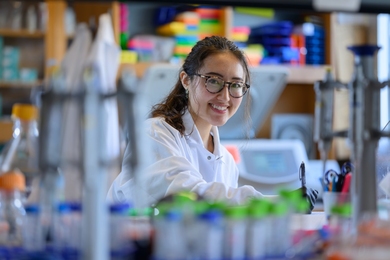 Photo of Katie Spivakovsky sitting at a lab bench, holding a pen and surrounded by lab equipment.