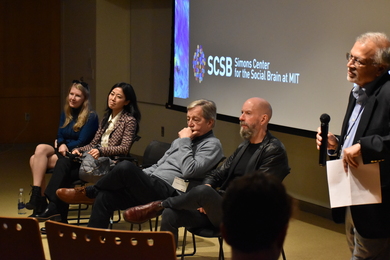 Ev Fedorenko, Gloria Choi, Charles Nelson, and Earl Miller sit in front of a screen with the logo of the Simons Center for the Social Brain. Mriganka Sur stands nearby, holding a microphone