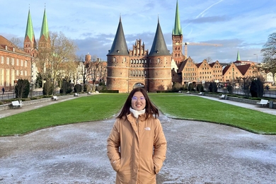 Crystal Liang, wearing a winter jacket, stands in front of a German castle with round towers and high, pointed spires