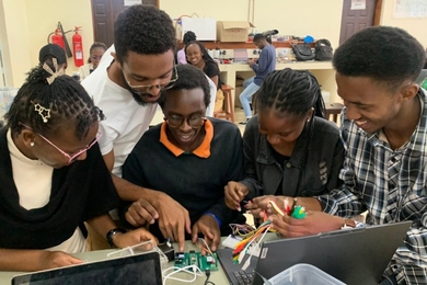 Five students gather around a table with an open laptop and parts needed to build a medical device