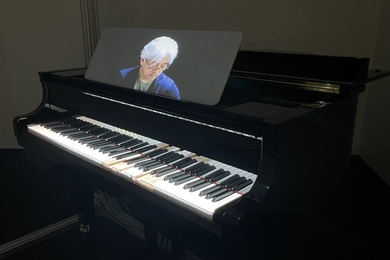 A spotlit piano in a darkened room with some keys depressed, and a screen showing a man looking down at the keyboard while playing