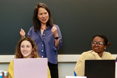 Lily Tsai stands behind Leela Fredlund and Karenna Caton, who are seated at a table with open laptops.