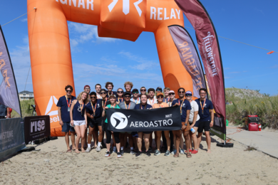 22 smiling teammates stand under a large orange inflatable arch on sandy terrain, holding an "AeroAstro MIT" banner
