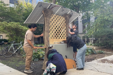 Four students installing solar-powered charging station comprising a metal electronics cabinet, two tree trunk-like pillars, wooden lattice, and four solar panels