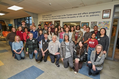 About three dozen casually dressed, smiling people pose in front of a wall bearing a mission statement that reads, “TASK feeds those who are hungry in the Trenton area and offers programs to encourage self-sufficiency and improve the quality of life of its patrons."
