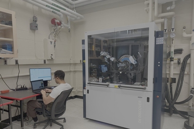 A man with his back to the camera works at a laptop next to lab equipment in a glass-and-steel cabinet