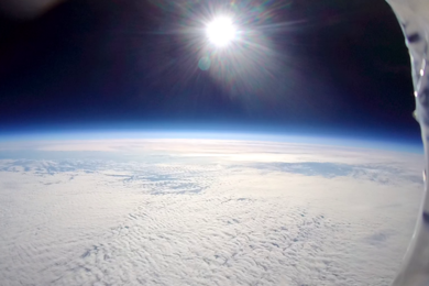 View of Earth's curvature from the stratosphere, with clouds below and the sun shining in a dark blue sky.