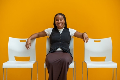 Olivia Honeycutt, a black woman with braids, poses seated on a white chair between two other white chairs before a golden yellow wall