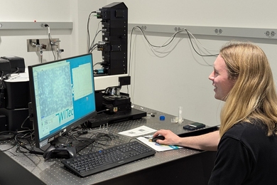 A person operating a computer mouse sits in front of a screen near an advanced microscope