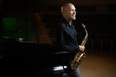 Miguel Zenón leans against piano and holds the saxophone in a dark music room.