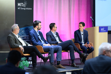 Four people sit in chairs on a stage indoors. One talks while the others look at him.