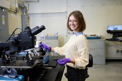Camille Cunin stands at a laboratory worktable using a microscope. She wears purple nitrile gloves and smiles broadly at the camera.