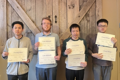Four students pose in front of a pair of rustic wooden doors. Each is holding up either one or two certificates bearing his name.