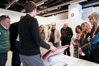 About a dozen students and faculty stand around a table at the MIT Museum as a presenter opens a large archival book