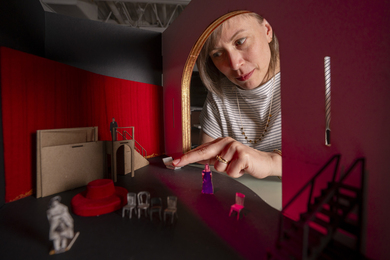 Sara Brown’s hand reaches through a tiny door into a miniature model of a red room with paper people and chairs.