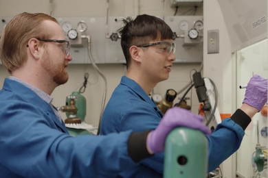 Glen Junor and Fang-Yu Kuo, wearing blue lab coats and safety glasses, work in a wet lab