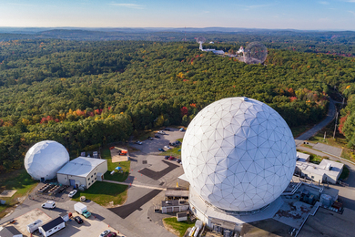 A large, white geodesic dome surrounded by low buildings, a smaller white dome, and a large forested area holding two large radio dishes