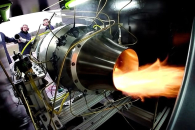 A jet engine producing waves of flame is seen from inside a test chamber. Two researchers stand at the front of the chamber.
