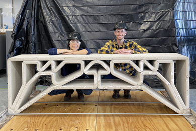 Hajin Kim-Tackowiak and Zane Schemmer, in hard hats and safety glasses, kneel behind a concrete bridge about the size of a park bench