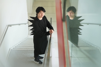 Pat Pataranutaporn stands on a staircase wearing a black coat with dramatic fabric spikes on his back, his reflection visible in a glass panel beside him.