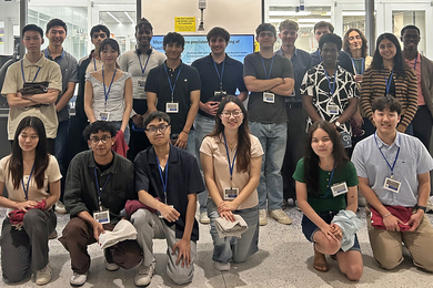 23 students wearing lanyards pose in a hallway with windows showing inside a clean lab