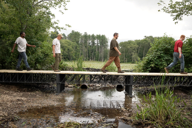 Four people walk across a 3-D printed bridge in a wetland.